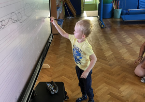 Dzieci siedzą na podłodze, a chłopiec rysuje na tablicy interaktywnej.