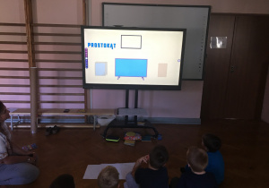 Dzieci siedzą na podłodze i oglądają film edukacyjny o figurach geometrycznych.