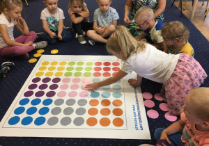 Dzieci siedzą na dywanie i układają kolorowe krążki na macie do kodowania.