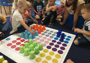 Dzieci siedzą na dywanie i układają kolorowe kueczki na macie do kodowania.