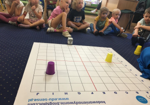 Dzieci siedzą na dywanie i słuchają o genibocie.