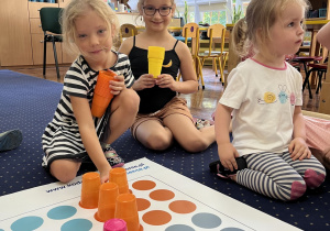 Dzieci siedzą na dywanie i układają kolorowe kubeczki na macie.