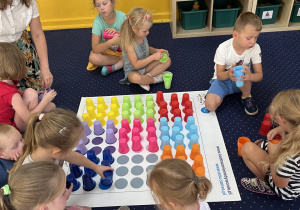 Dzieci siedzą na dywanie i układają kolorowe kubeczki na macie.