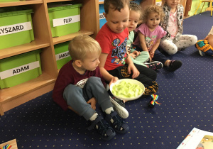 Dzieci siedzą na dywanie i przekazują sobie talerz z warzywami.