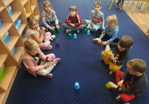 Dzieci siedzą na podłodze i trzymają kolorowe kubeczki.