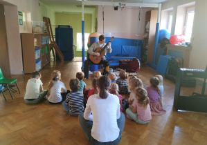 chłopiec gra na gitarze, dzieci siedzą na podłodze na sali gimnastycznej, słuchają muzyki