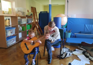 dziewczynka trzyma gitarę, starszy chłopiec pokazuje jak prawidłowo chwytać gitarę i na niej grać