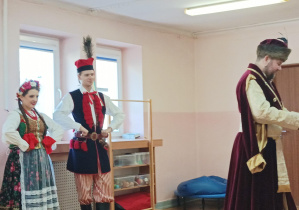 kobieta i mężczyzna ubrani w stroje krakowskie, stoją na sali gimnastycznej, obok widać mężczyznę w stroju szlacheckim