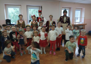 grupa dzieci pozuje do zdjęcia, za nimi widać grupę osób ubranych w stroje krakowskie i szlacheckie