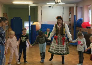 dzieci na sali gimnastycznej tańczą krakowiaka wraz z osobami ubranymi w stroje ludowego