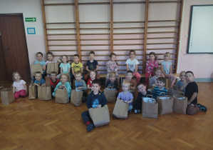 grupa dzieci siedzi na sali gimnastycznej, każde dziecko trzyma papierową torbę z nagrodami