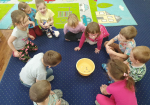 Dzieci siedzą w kole na dywanie i obserwują monety w misce.