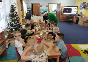 dzieci przy stolikach wykonują świątecznego skrzata, pomagają im dwie panie. Jedna w stroju elfa, druga w mikołajowej czapce.