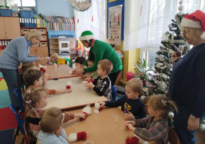 dzieci przy stolikach wykonują świątecznego skrzata, pomagają im dwie panie. Jedna w stroju elfa, druga w mikołajowej czapce.
