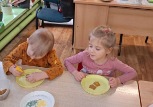 dwoje dzieci siedzi przy stoliku, na talerzach mają ciastka, które dekorują