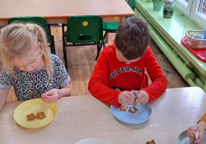 dwoje dzieci siedzi przy stoliku, na talerzach mają ciastka, które dekorują
