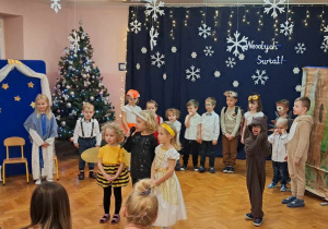 dzieci w przebraniach zwierząt wystepują na sali gimnastycznej, recytują wiersze do mikrofonu, śpiewają piosenki, tańczą. W tle napis "Wesołych Świąt".