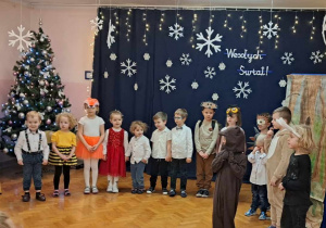 dzieci w przebraniach zwierząt wystepują na sali gimnastycznej, recytują wiersze do mikrofonu, śpiewają piosenki, tańczą. W tle napis "Wesołych Świąt".
