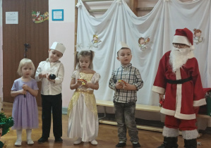 dzieci na sali gimnastycznej występują na przedstawieniu choinkowym, chłopiec w przebraniu Mikołaja, dziewczynki w sukienkach, chłopcy w eleganckich strojach