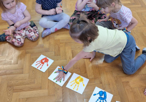 dzieci siedzą na podłodze na sali gimnastycznej, przed nimi leżą karki ze śladem dłoni w różnych kolorach