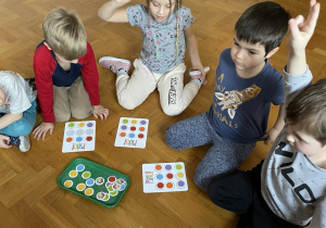 dzieci siedzą na podłodze na sali gimnastycznej, przed nimi leżą kartki z kolorowym wzorem