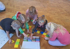dzieci siedzą na podłodze na sali gimnastycznej, przed nimi duża mata do kodowania i kolorowe karteczki