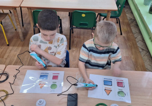 dwóch chłopców przy stoliku odrysowuje kształt figur geometrycznych przy pomocy długopisu 3D