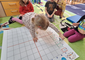 dzieci siedzą w sali na dywanie, przed nimi duża mata do kodowania