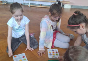 dzieci siedzą na podłodze na sali gimnastycznej, układają kolorowe kartoniki