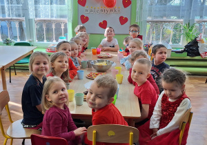 dzieci w czerwonych strojach siedzą przy stolikach, w tle walentynkowa dekoracja