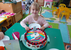 Dziewczynka siedzi przy stole, przed nią znajduje się tort.