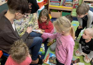 Dzieci siedzą na dywanie. Dziewczynka zaznacza odpowiedź w książce.