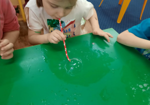 Dzieci siedzą przy stole i bawią się roztworem mydlanym.