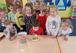dzieci z grupy I stoją za stołem, na którym widać symboliczny tort, chłopiec dmucha świeczkę