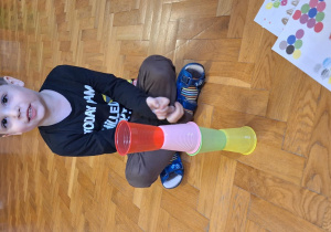 Chłopiec siedzi na podłodze, a przed nim stoją ułożone kolorowe kubeczki.