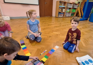Dzieci siedzą na podłodze i układają kolorowe karteczki.
