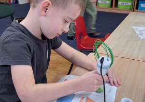 Chłopiec siedzi przy stoliku i wykonuje zadanie wykorzystując długopis 3D.