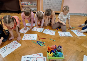 Dzieci siedzą na podłodze, przed nimi leżą kartki z napisem "LOVE".