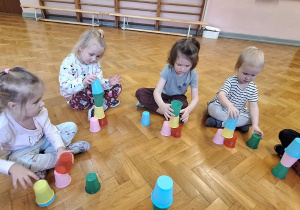 Dzieci siedzą na podłodze i układają kolorowe kubeczki.