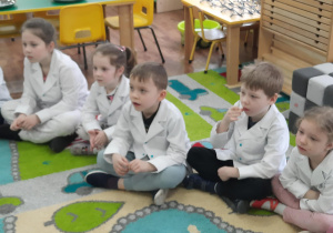 Dzieci siedzą w kole w białych fartuszkach laboratoryjnych.