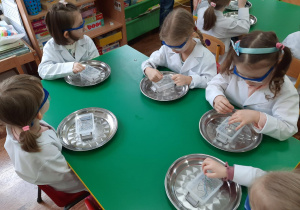 Dzieci siedzą przy stolikach i oglądają zgromadzone w pojemniku różne materiały.
