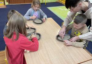 Dzieci siedzą przy stolikach i piszą na tackach z piaskiem.