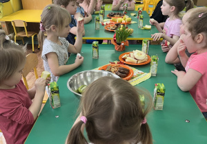 Dzieci siedzą przy stole i zjadają owoce i słodkości popijając soczkami.