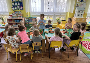 Dzieci siedzą przy stole i zjadają owoce i słodkości popijając soczkami.