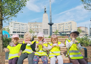 grupa dzieci w żółtych kamizelkach siedzi na ławce na Placu Wolności