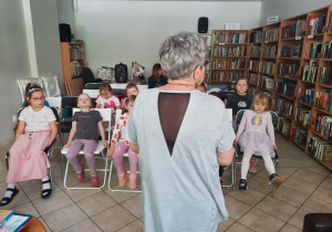 Dzieci siedzą na krzesłach i słuchają pani bibliotekarki.