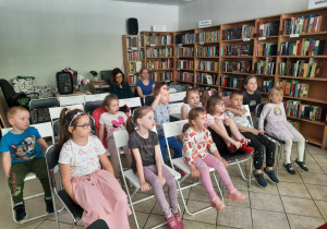 Dzieci siedzą na krzesłach i słuchają pani bibliotekarki.