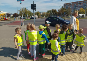 dzieci pod opieką nauczycielki, w kamizelkach odblaskowych, spacerują po chodniku