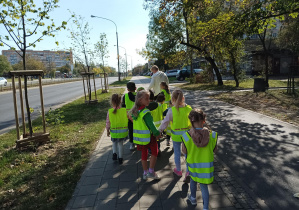 dzieci pod opieką nauczycielki, w kamizelkach odblaskowych, spacerują po chodniku