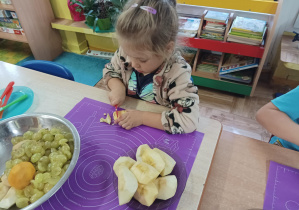 dziewczynka siedzi przy stoliku i kroi owoce na sałatkę owocową.